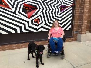 Karin Willison sitting in my wheelchair outside in front of a zigzag mural.