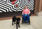 Karin Willison with my service dog sitting in my wheelchair outside in front of a zigzag mural.