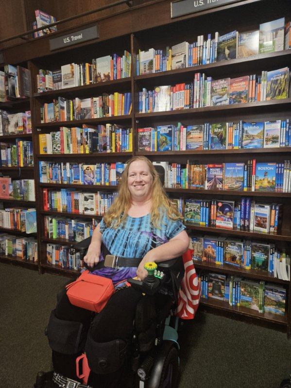 Karin Willison sitting in her power wheelchair with a wall of travel books in the background.